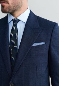 Marineblauwe geruite blazer met notch-lapels, gecombineerd met een lichtblauw overhemd en een gestippelde stropdas met marineblauwe en lichtgrijze cirkels.