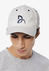 Gorra blanca de algodón con un logo bordado en azul marino, seis orificios de ventilación y visera curvada; llevada por un modelo con una camisa gris.