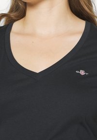 Černé bavlněné tričko s výstřihem do V. Na levé části hrudi se nachází malé vyšívané logo. Hladký materiál a přiléhavý střih.