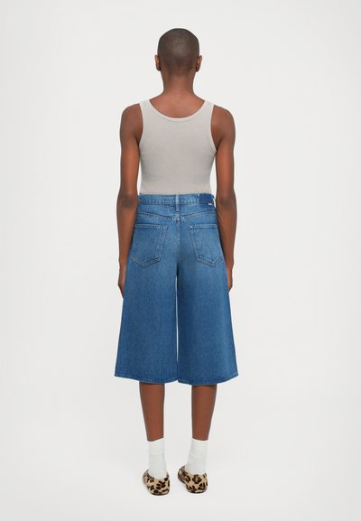 Person with short hair wearing a sleeveless beige top, wide blue denim culottes, white socks, and leopard-print slip-on shoes, facing away.