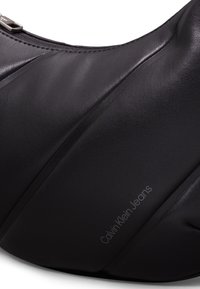 Černá taška z umělé kůže s hladkým povrchem a zakřiveným designem. Disponuje jemným prošíváním a embosovaným logem "Calvin Klein Jeans".