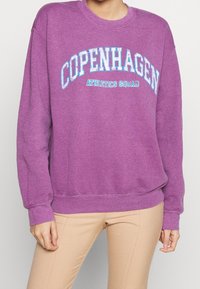 Fialová mikina s potiskem "COPENHAGEN ATHLETICS SQUAD" v modré barvě, dlouhé rukávy, kulatý výstřih a měkká textura.