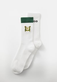 Calcetines blancos con puños acanalados verdes. Un calcetín presenta un logo bordado en oro, mientras que el otro tiene "HUGO" impreso en negro.