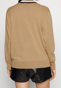 Femme portant un pull couleur camel avec un col rayé et un short en vinyle noir brillant, vue de dos sur un fond uni.