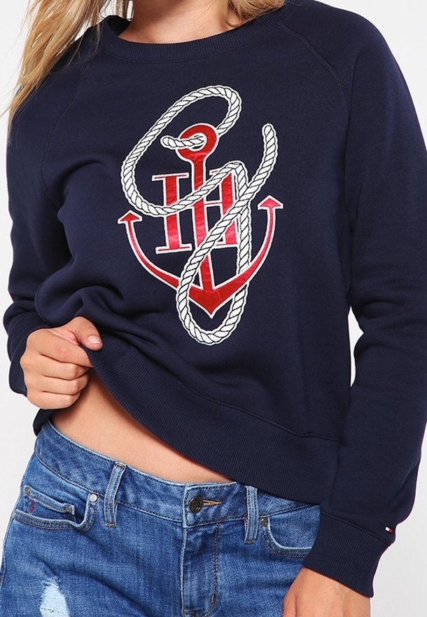 Felpa blu navy con grafica di ancora bianca e rossa. Presenta polsini e orlo a costine, tessuto morbido e vestibilità comoda. Indossata con jeans azzurri.