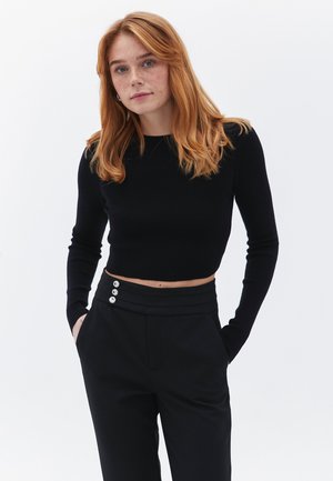 Jersey negro corto con mangas largas combinado con pantalones negros de cintura alta que presentan cuatro botones decorativos en la cinturilla.