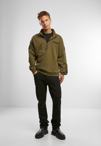 Pullover in pile verde oliva con colletto zip, due tasche sul petto e polsini a costine. Abbinato a jeans neri aderenti e stivali neri.