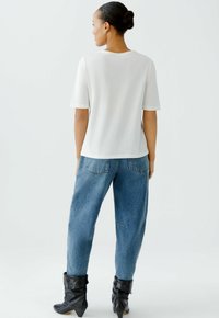 Frau mit einem Dutt, die ein weißes Kurzarmhemd, eine blaue Jeans und schwarze, lässige Stiefeletten trägt, steht mit dem Rücken zu einem schlichten Hintergrund.