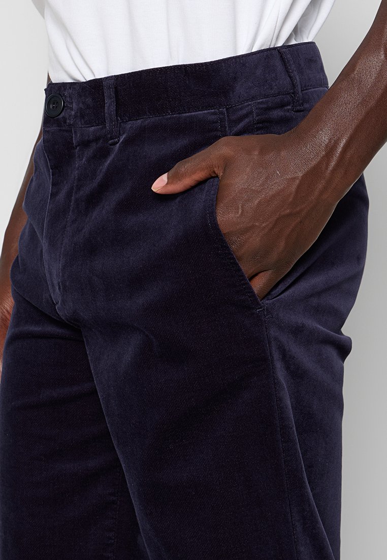 Donkerblauwe ribfluwelen broek gedragen door een persoon met de hand in de rechtervoorzak, gecombineerd met een ingestopt wit overhemd.