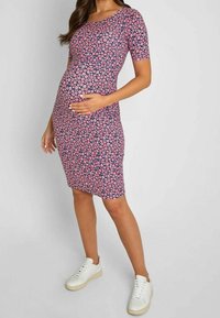 Femme enceinte portant une robe rose fleurie longueur genoux et des baskets blanches, tenant son ventre d'une main.