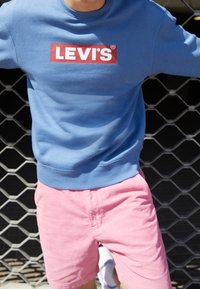 Felpa blu con logo rosso Levi's, abbinata a pantaloni corti rosa chiaro. I materiali sembrano morbidi, con una vestibilità comoda e un design informale.