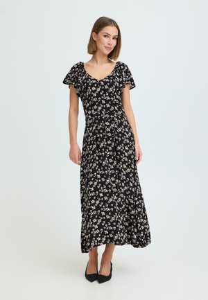 Kvinde stående iført en sort blomstret midi-kjole med flagrende ærmer og sorte hæle mod en ensfarvet lys baggrund.