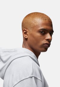 Lysegrå hættetrøje med snor og synlige sømme, iført af en model med kort, cropped gyldent hår, mod en hvid baggrund.