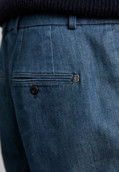Pantalonii din denim bleu închis au un buzunar pe spate cu închidere cu nasture și un mic detaliu cu fermoar deasupra. Tesătura are o textură netedă.
