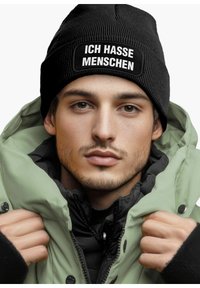 Black ribbed beanie with white text "ICH HASSE MENSCHEN," worn under a light green puffer jacket, with a black inner layer and visible buttons.