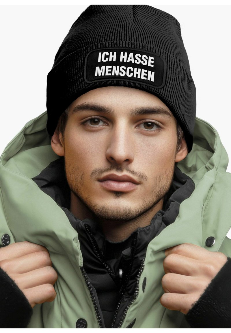 Black ribbed beanie with white text "ICH HASSE MENSCHEN," worn under a light green puffer jacket, with a black inner layer and visible buttons.