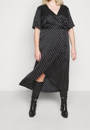 Robe noire à pois blancs, à manches courtes et avec une fente sur le côté. Associée à des bottes noires jusqu'aux genoux. Tissu lisse, design en v.