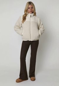 Veste en polaire crème avec fermeture éclair à l'avant, design à panneaux contrastants et poignets côtelés. Portée avec un pantalon évasé marron et des chaussures marron clair.