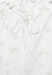 Tessuto bianco con elaborati ricami floreali argento, caratterizzato da una chiusura con cordino e una superficie liscia. Una cucitura decorativa delinea il design.