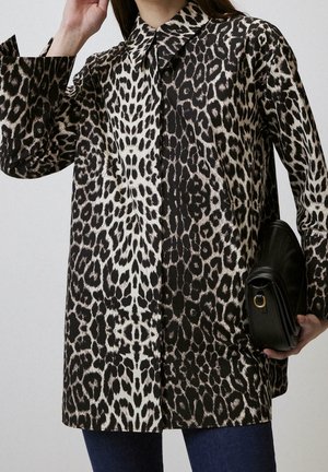 Mujer con camisa de manga larga con estampado de leopardo y patrón dividido en negro y blanco, sosteniendo un bolso de mano negro de cuero y vistiendo vaqueros azules.