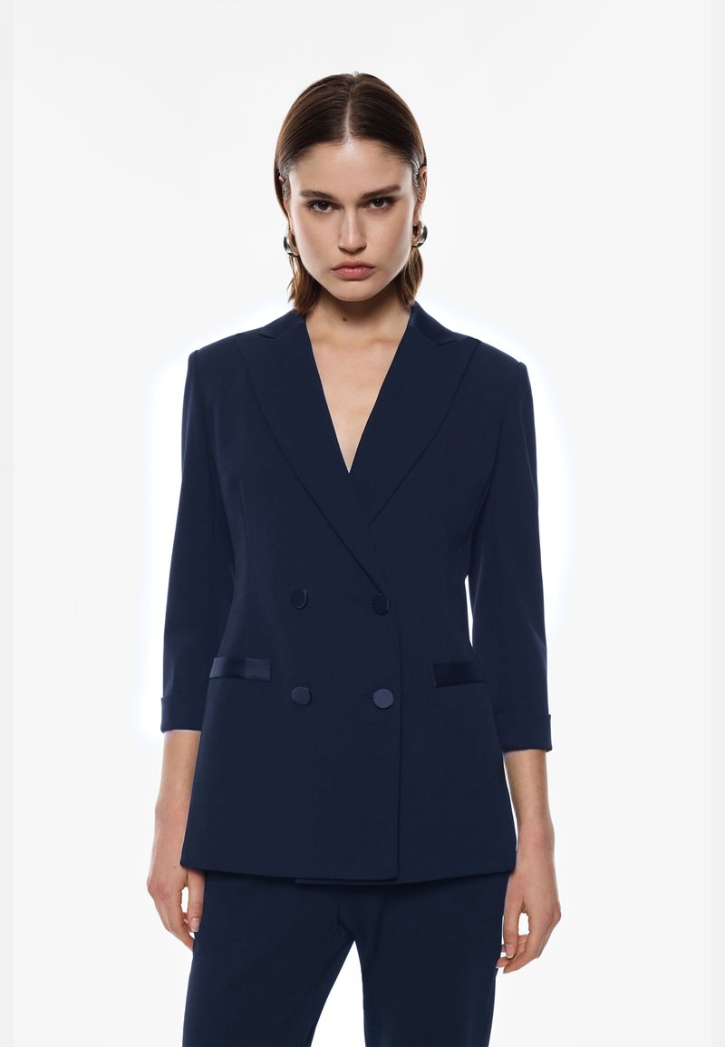 Blazer doppiopetto blu navy con scollatura a V profonda, maniche a tre quarti, dettagli in satin e reverse ampi; vestibilità sartoriale con design strutturato.