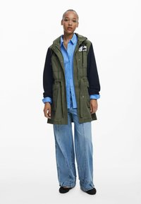 Olivgrüne Parka mit dunkelblauen Ärmeln, tailliertem Schnitt und Fronttaschen, über einem hellblauen Hemd und weit geschnittenen Jeans.