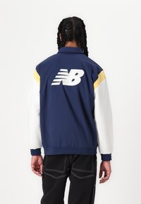 Veste légère marine avec des accents jaunes et blancs. Présente un grand logo blanc "NB" au dos et des poignets côtelés pour un look ajusté.