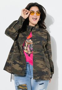 Giacca con stampa camouflage in verde scuro e marrone con colletto, bottoni a scatto e tasche laterali, indossata sopra una t-shirt grafica rosa e jeans strappati.