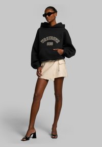 Black hooded sweatshirt with logo, cream shorts, and black block-heeled sandals. Smooth fabric and relaxed fit with front pocket.