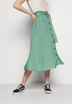 Mujer que lleva una falda midi verde de talle alto con botones delanteros y cinturón, top beige y zapatillas negras y blancas, posando.