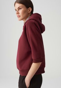 Hoodie corto bordeaux con cappuccio regolabile, maniche corte e orlo a coste. Tessuto liscio con dettagli delle cuciture sulle maniche.