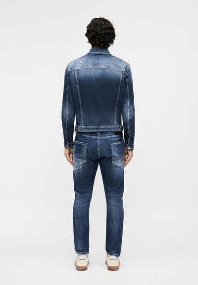 Homme portant une veste en denim bleu et un jean avec des détails délavés et usés, debout de dos devant un fond uni.