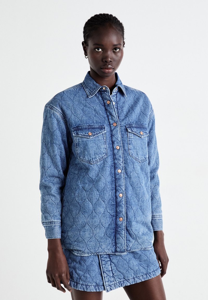 Jeans srajca v svetlo modri barvi z zloženim vzorcem, zapenjanjem s gumbi in dvema žepoma na prsih, kombinirana z ujemajočo se jeans krilom.