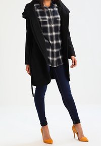 Svart lång kappa med huva, rutig skjorta i marinblått och vitt, mörkblå tajta jeans och gula högklackade skor. Släta texturer, rena linjer.