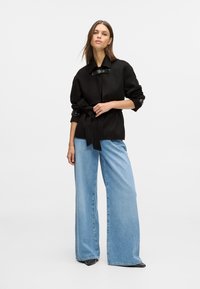 Czarna kurtka zapinana na pasek z wysokim kołnierzem, w połączeniu z jasnoniebieskimi dżinsami o szerokich nogawkach i spiczastymi czarnymi butami. Gładki materiał, minimalne detale.