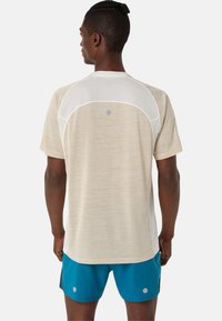 T-shirt de sport à manches courtes en beige clair avec un tissu texturé et des panneaux en mesh, doté d'un col rond et de détails en argent à l'arrière.