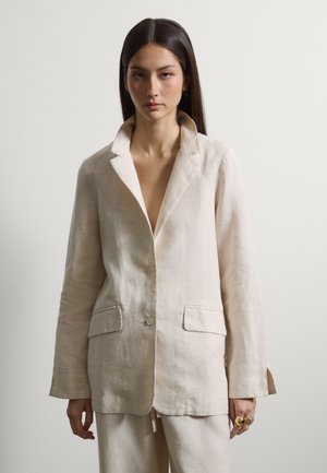 Mujer con cabello largo y oscuro, vistiendo un blazer de lino suelto color crema y pantalones a juego, de pie contra un fondo claro y liso.