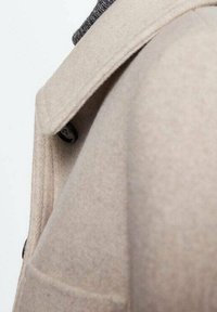 Beige uldfrakke med et struktureret design, der har en spids krave, to knapper og et gråt ribbet stof under.