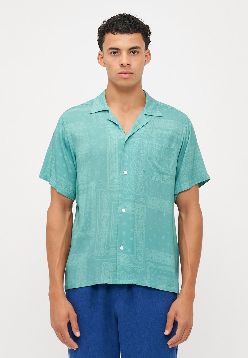 Camisa de manga curta em teal com padrões intrincados, um bolso no peito, fechamento com botões e um corte descontraído. O tecido parece leve e suave.