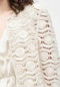 Λευκό δαντελένιο ύφασμα πλεκτό με κυκλικά και κυματιστά μοτίβα, διακοσμημένο με μικρές παγιέτες πάνω σε σατέν ένδυμα, φορεμένο από ένα άτομο με καστανά μαλλιά.