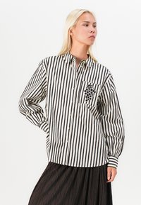 Camicia a righe bianche e nere con taschino sul petto decorato da un motivo a grappolo d'uva. Maniche lunghe e vestibilità morbida.