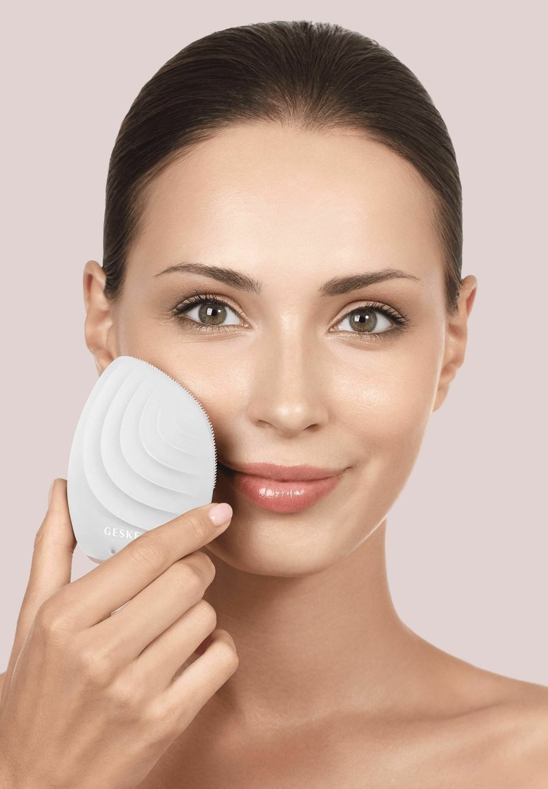 Woman with smooth skin holds a white, textured facial cleansing brush to her right cheek against a plain light background.