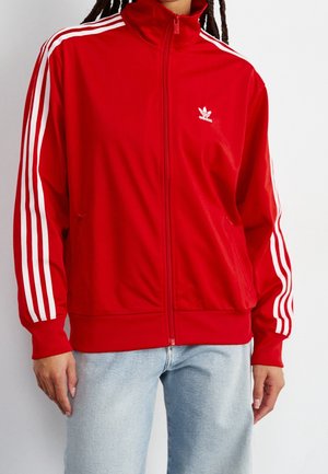 Punainen Adidas-verryttelytakki valkoisilla raidoilla hihoissa ja pienellä logolla rinnassa, käytössä vaaleansinisten farkkujen kanssa.