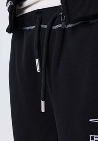 Primer plano de pantalones de chándal negros con costuras blancas y cordones negros con puntas plateadas metálicas, acompañados de una chaqueta negra parcialmente visible.