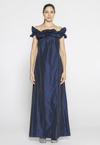 Granatowa sukienka off-the-shoulder z falbaniastym dekoltem, zwiewnym materiałem i długą sylwetką. Gładka tekstura, zestawiona z niebieskimi sandałami na odkrytej stópce.