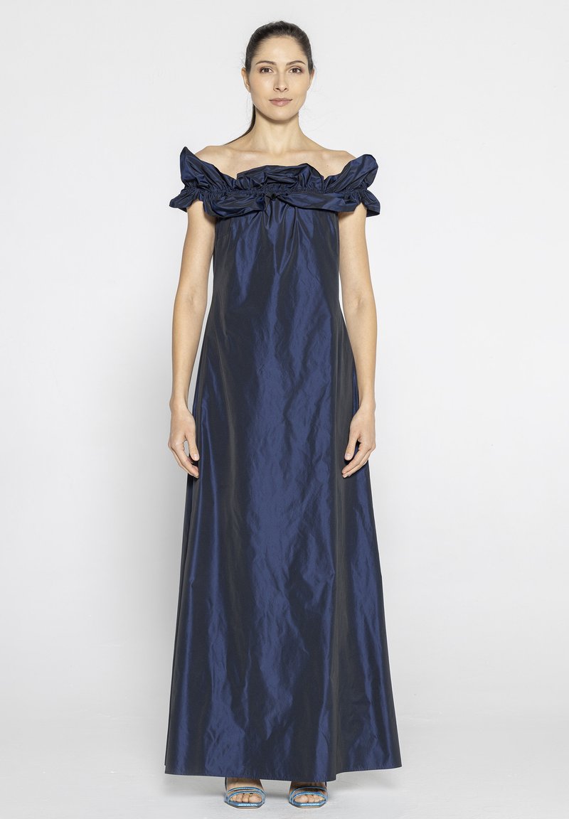 Granatowa sukienka off-the-shoulder z falbaniastym dekoltem, zwiewnym materiałem i długą sylwetką. Gładka tekstura, zestawiona z niebieskimi sandałami na odkrytej stópce.