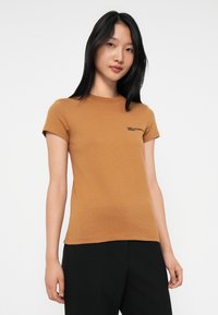 Jonge vrouw met lang zwart haar in een strakke bruine t-shirt met korte mouwen en zwarte broek, staand tegen een witte achtergrond.