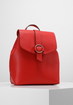 Sac à dos en cuir rouge avec une fermeture à rabat et un accent en métal circulaire, équipé d'une poignée robuste. Texture lisse et forme structurée.