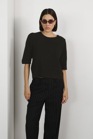 Black knit sweater with short sleeves, paired with black pinstriped trousers. Model wears oval sunglasses and simple hoop earrings.