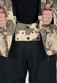 Kameliantekstuuri takki beežide ja pruunide toonidega, nööpidega, kantud mustade riiete peal. Suletud käed ja rõiva detailid.